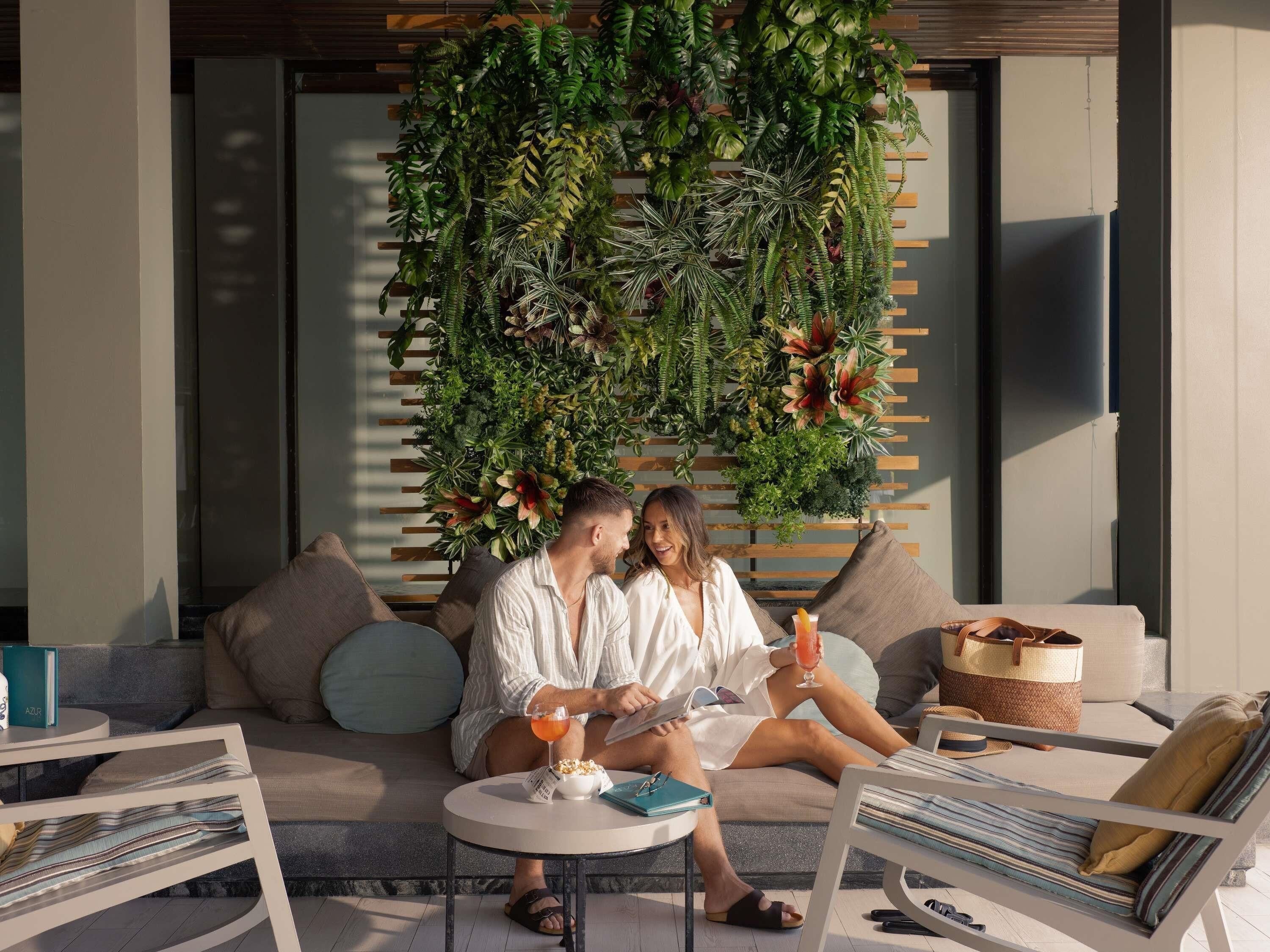 A couple sitting on a sofa with a lush green wall behind them, a small round table with a drink, and a magazine.