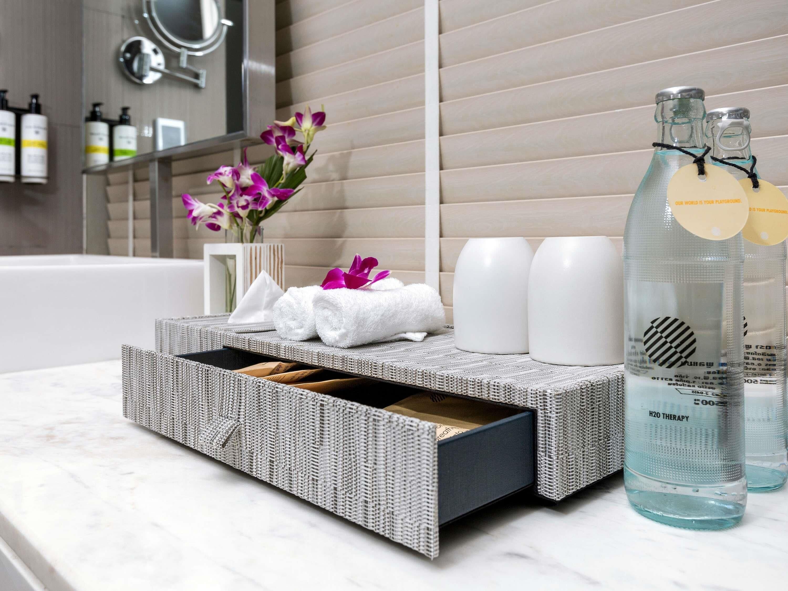 A bathroom vanity with a woven basket, white towels, a vase with purple flowers, and two bottles labeled "H2O THERAPY".