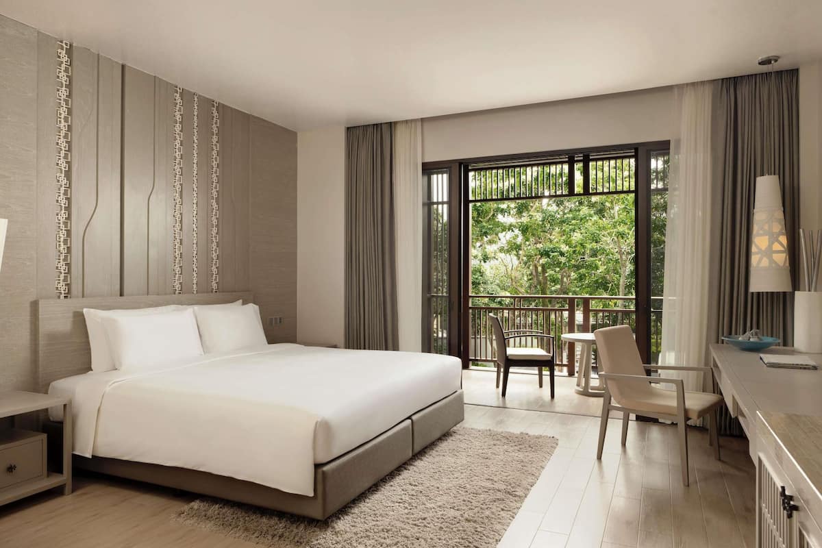 Quarto de hotel moderno com uma cama grande, escrivaninha, cadeira e varanda com vista para árvores.