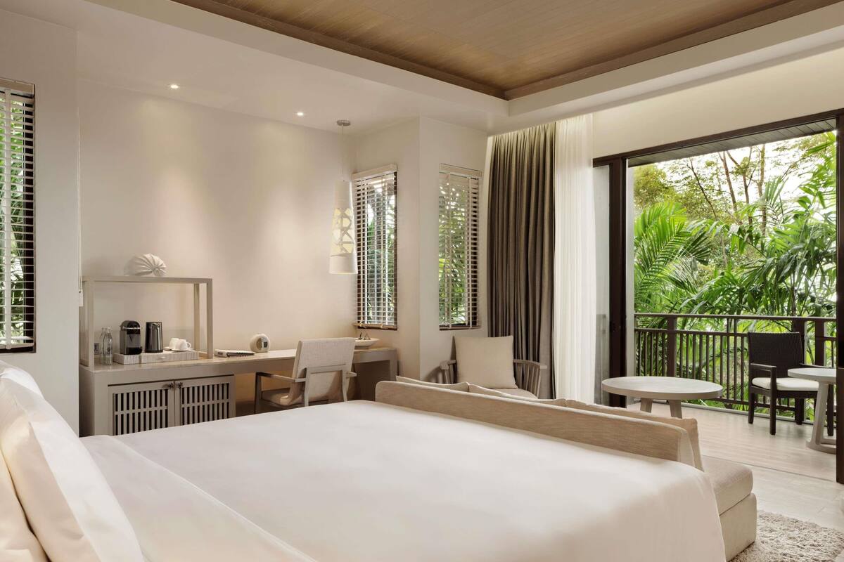 Quarto de hotel com uma cama grande, uma escrivaninha com cadeira, uma mesinha com cadeiras e uma varanda com vista para o verde.