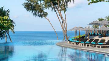 A pool with lounge chairs and umbrellas, palm trees, and a view of the ocean.