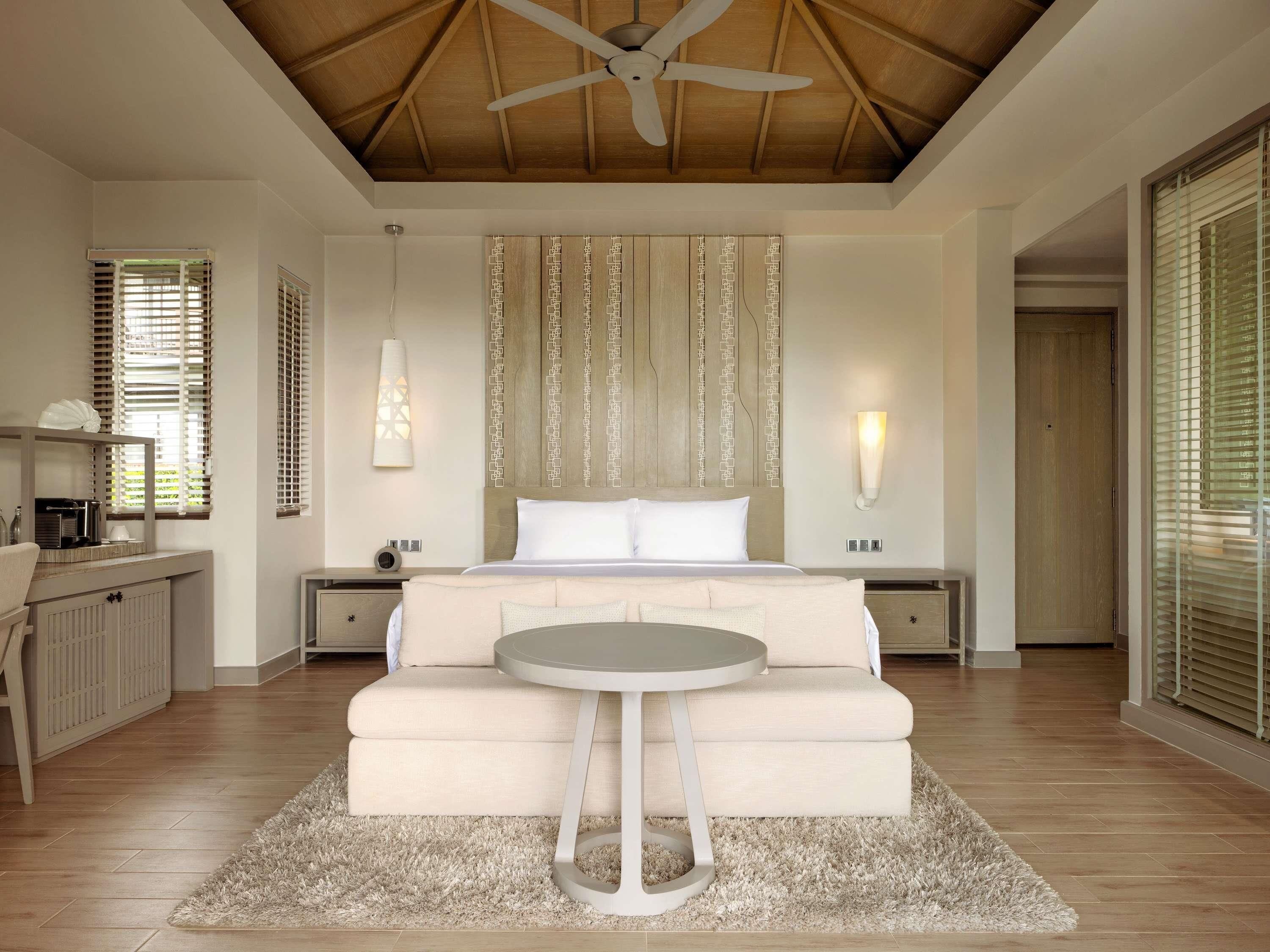A bedroom with a bed, bedside tables, a round table, a ceiling fan, and a large window with blinds.