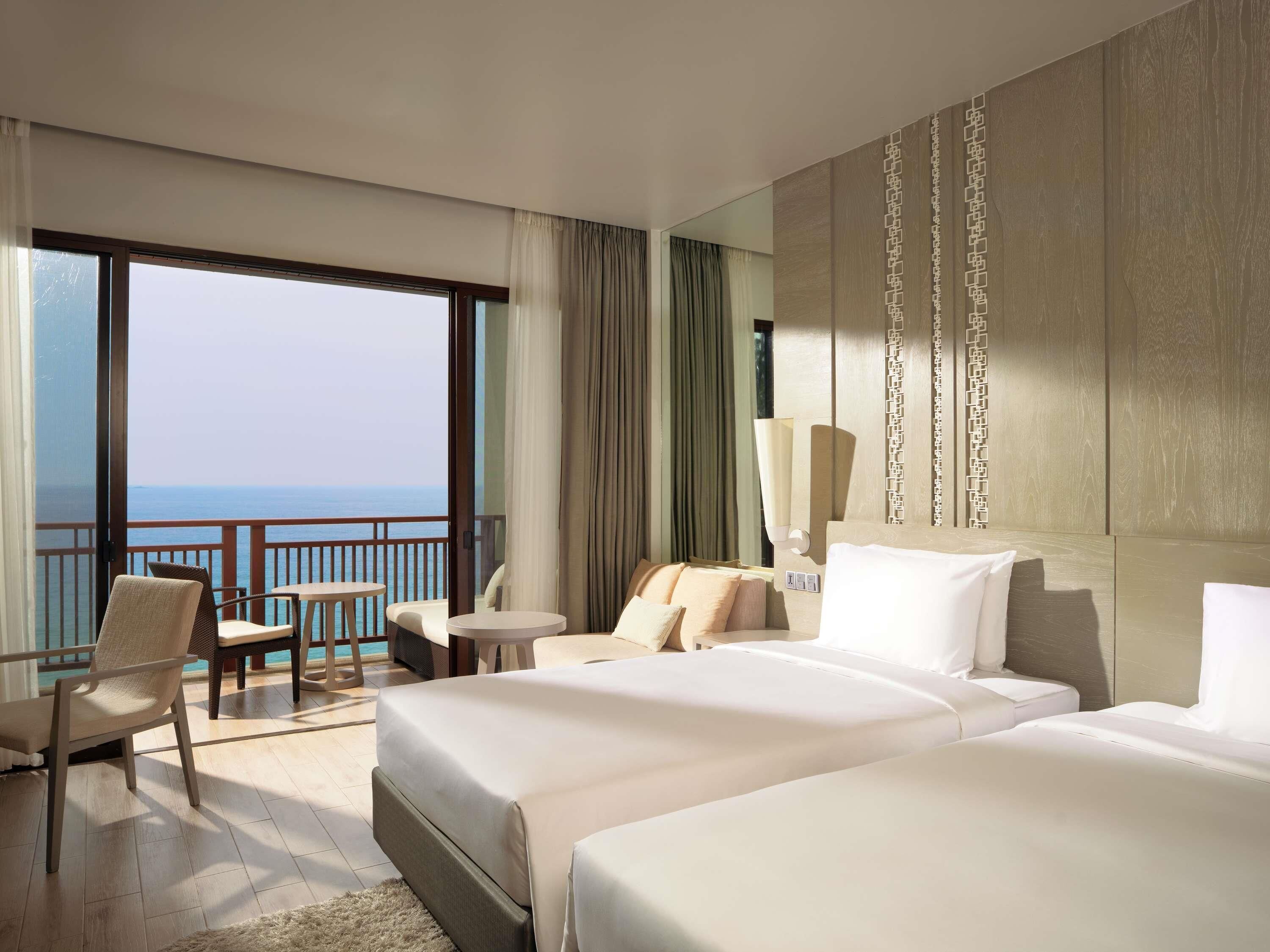 A hotel room with two beds, a balcony with a view of the ocean, a small table, and a chair.