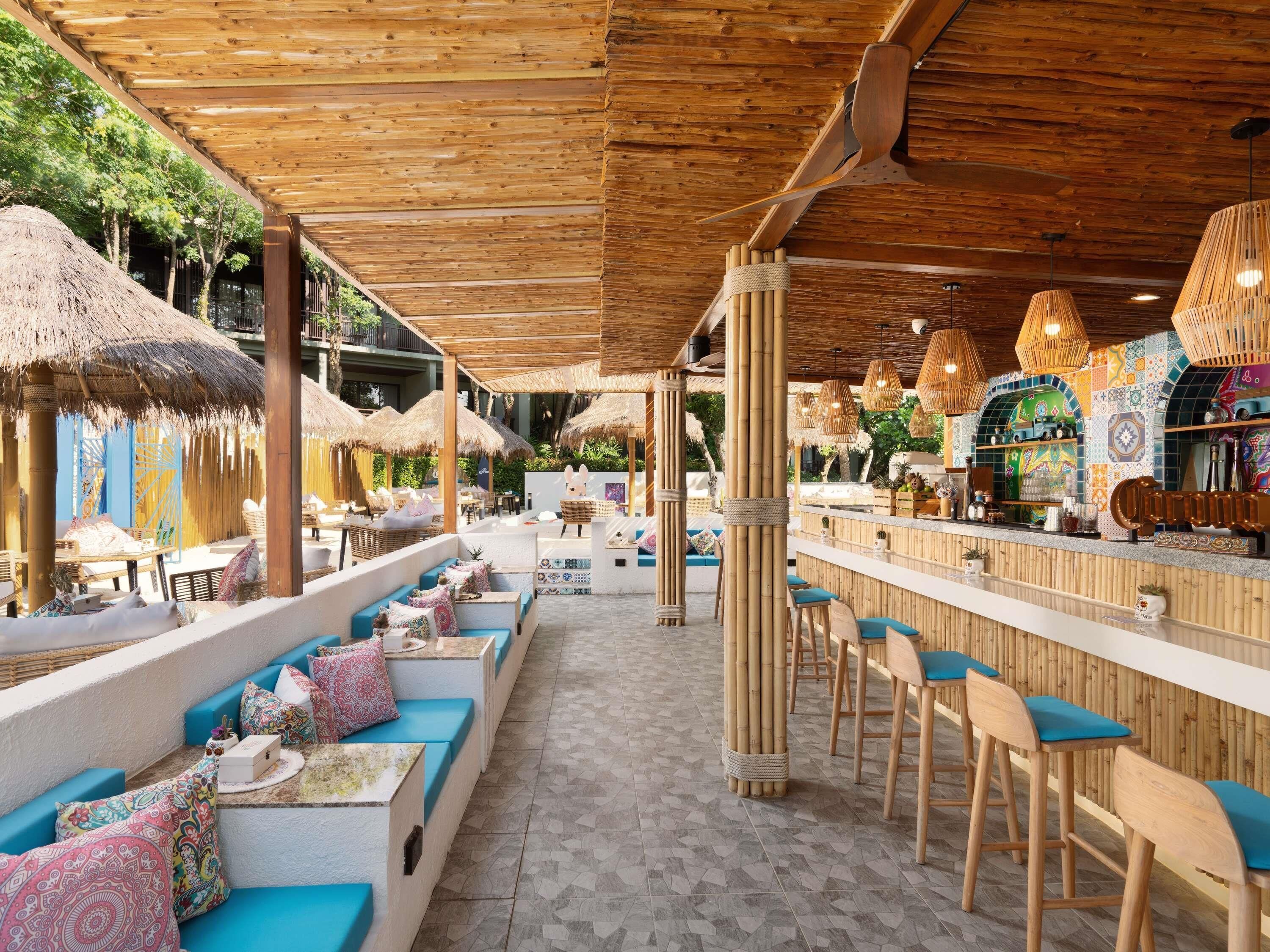 Un espace extérieur aménagé avec des parasols en chaume, du mobilier en bois et un bar dont le mur arrière est décoré de carreaux colorés.