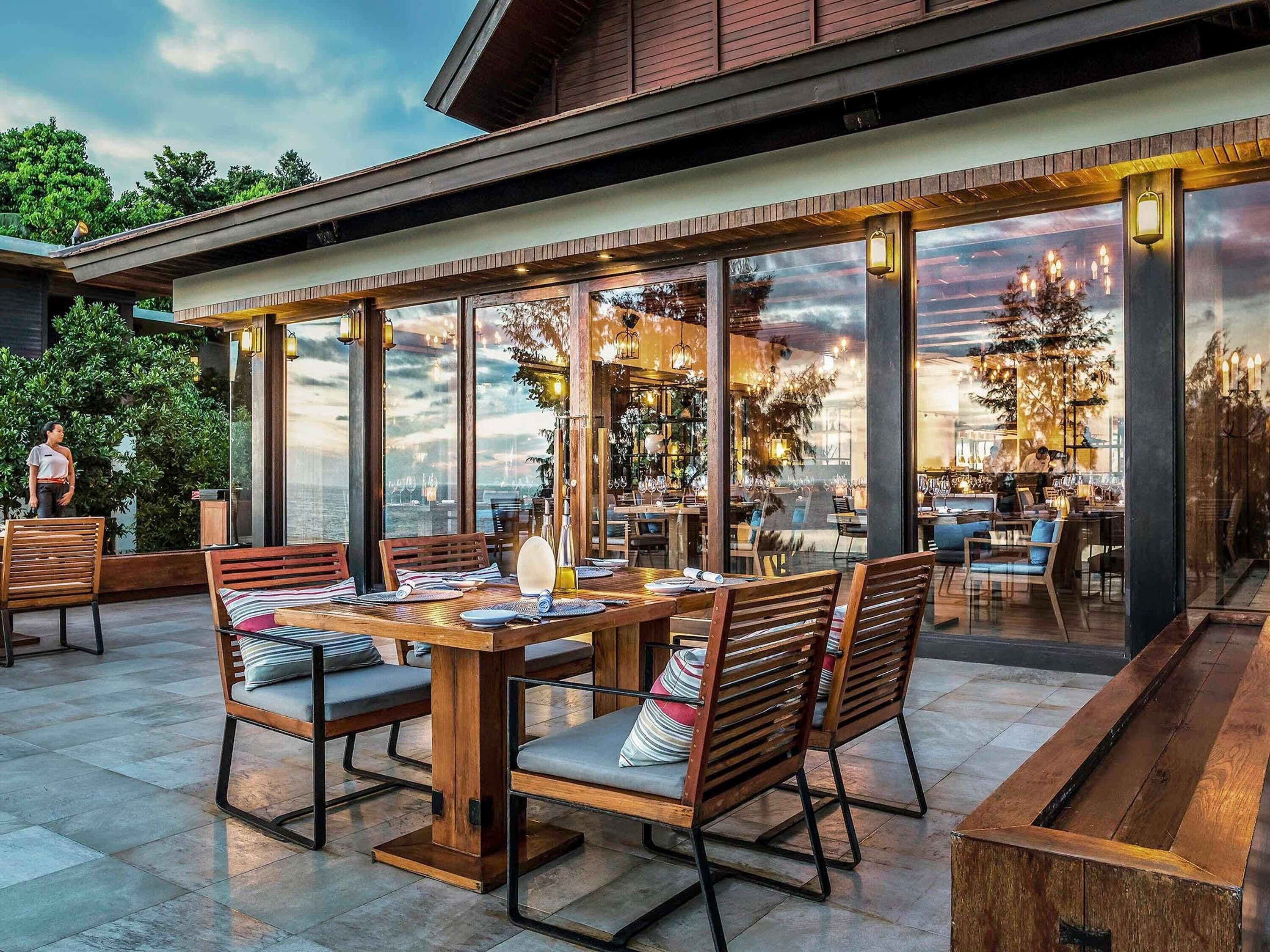 An outdoor dining area with wooden tables and chairs, large windows, and a view of the sky.