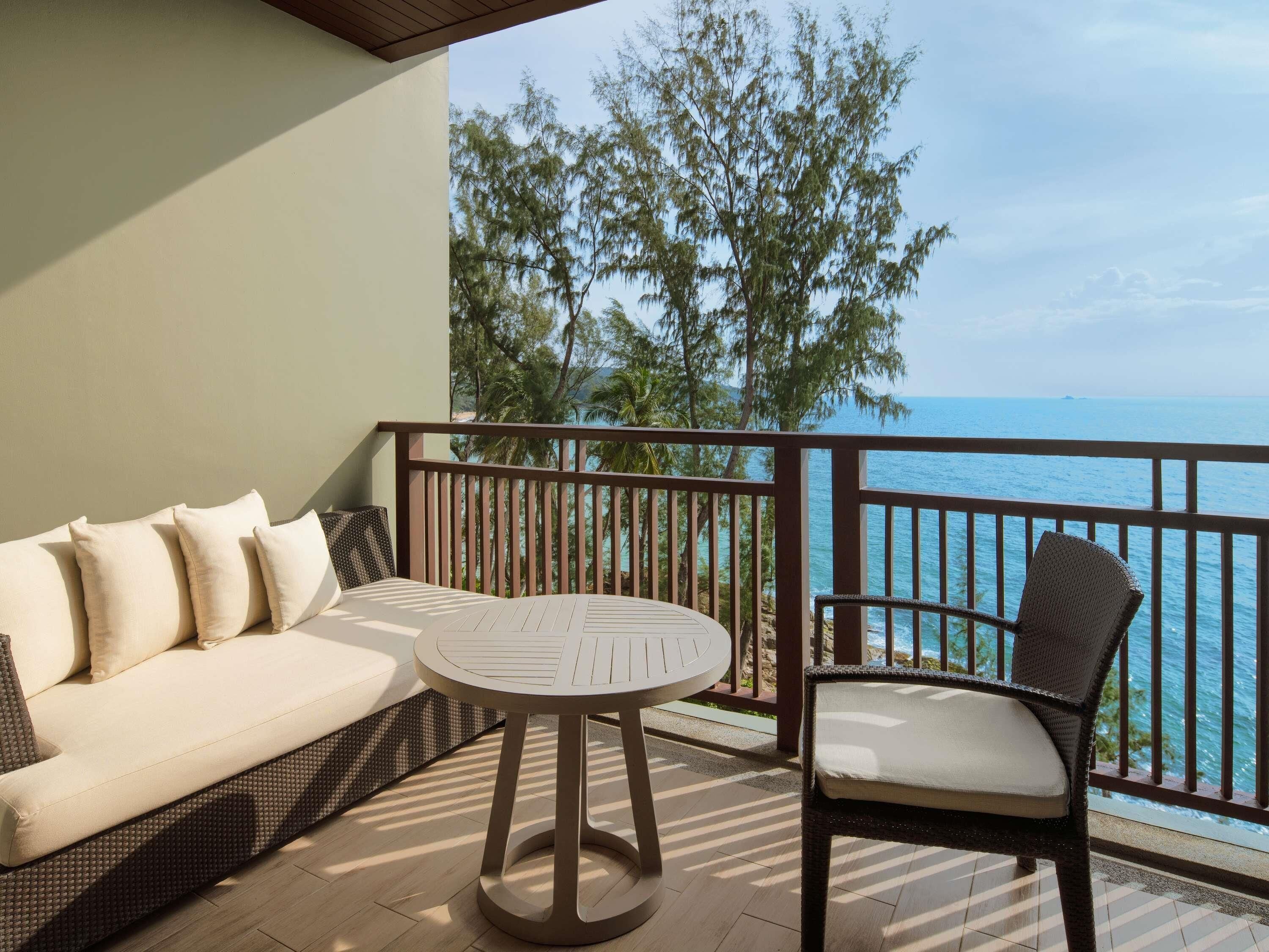 A balcony with a sofa, a round table, and a chair, overlooking a body of water and trees.