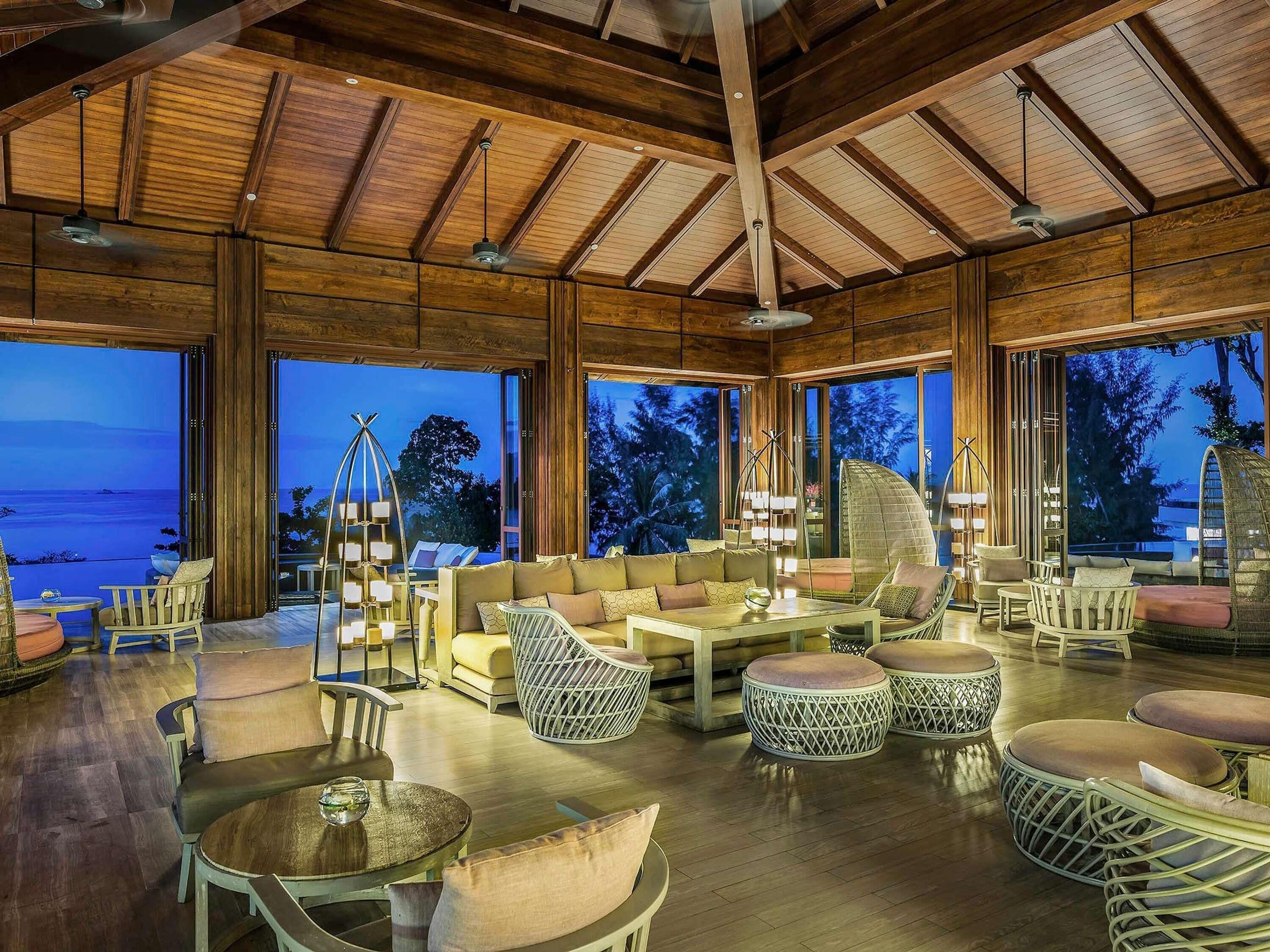 A spacious indoor area with wooden beams, large windows, and a variety of seating arrangements including sofas, chairs, and round tables.