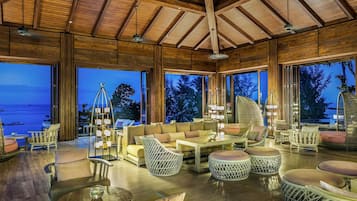 A spacious indoor area with wooden beams, large windows, and a variety of seating arrangements including sofas, chairs, and round tables.