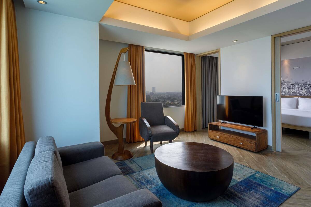 Ein modernes Hotelzimmer mit Sofa, Sessel, rundem Couchtisch, Fernseher und Blick auf die Stadt.