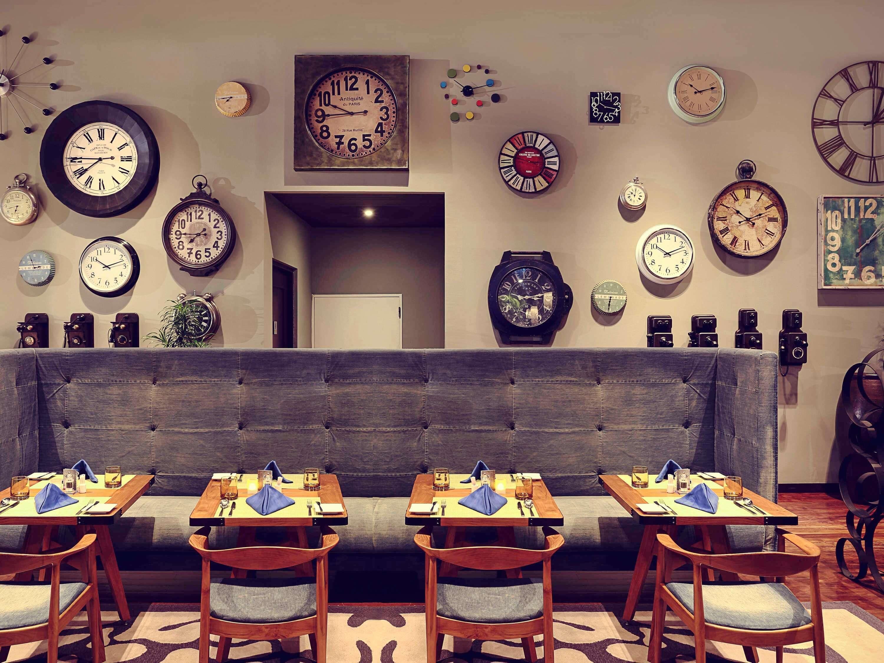 A dining area with multiple tables and chairs, a variety of wall clocks, and a neutral-toned sofa.