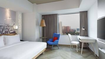 Quarto de hotel com cama, escrivaninha, cadeira, luminária, janela grande e vista para a cidade.