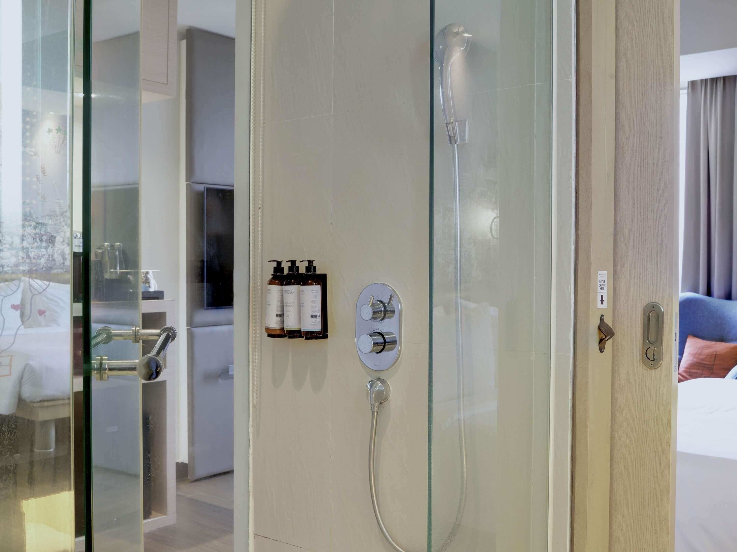 Habitación de hotel con cabina de ducha de vidrio, panel de control para la temperatura y el caudal del agua, y un pequeño estante con artículos de aseo.