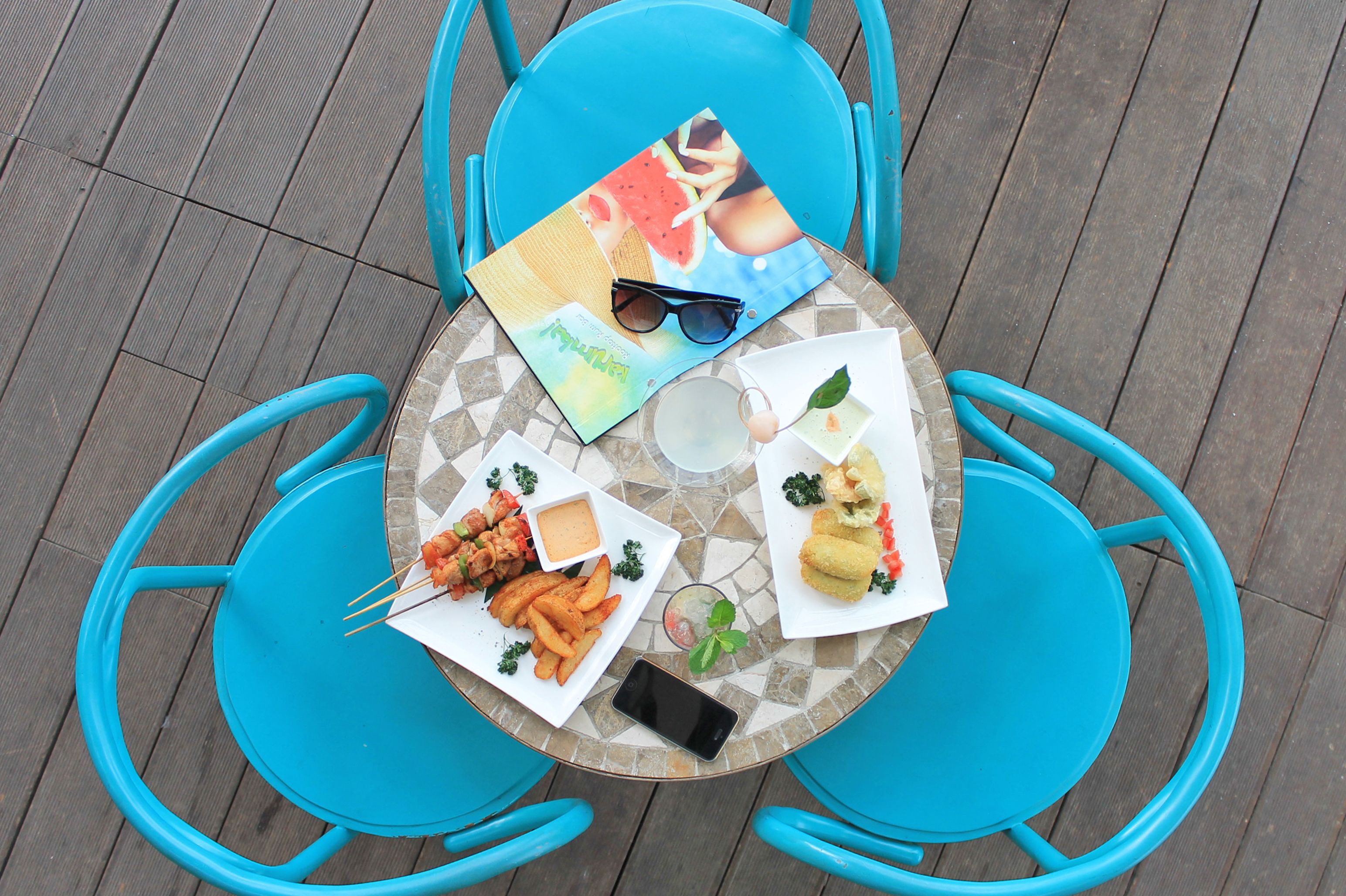 A round table with a mosaic pattern, surrounded by four blue chairs. On the table, there are plates of food including fries, a sandwich, and a dessert. A smartphone and a magazine are also visible.
