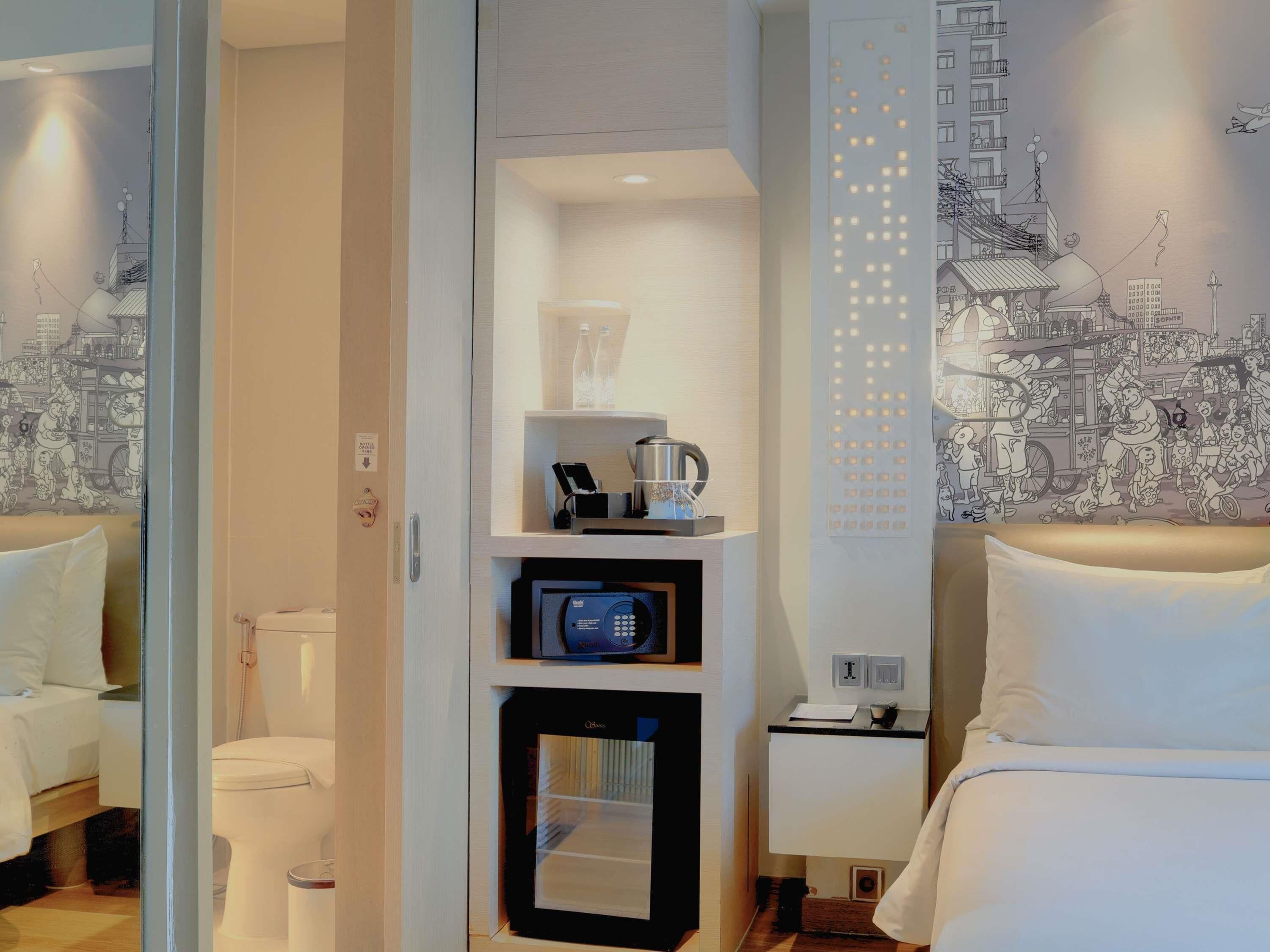 A hotel room with a bed, a bedside table with a lamp, a wall-mounted TV, a minibar, and a bathroom visible through an open door.