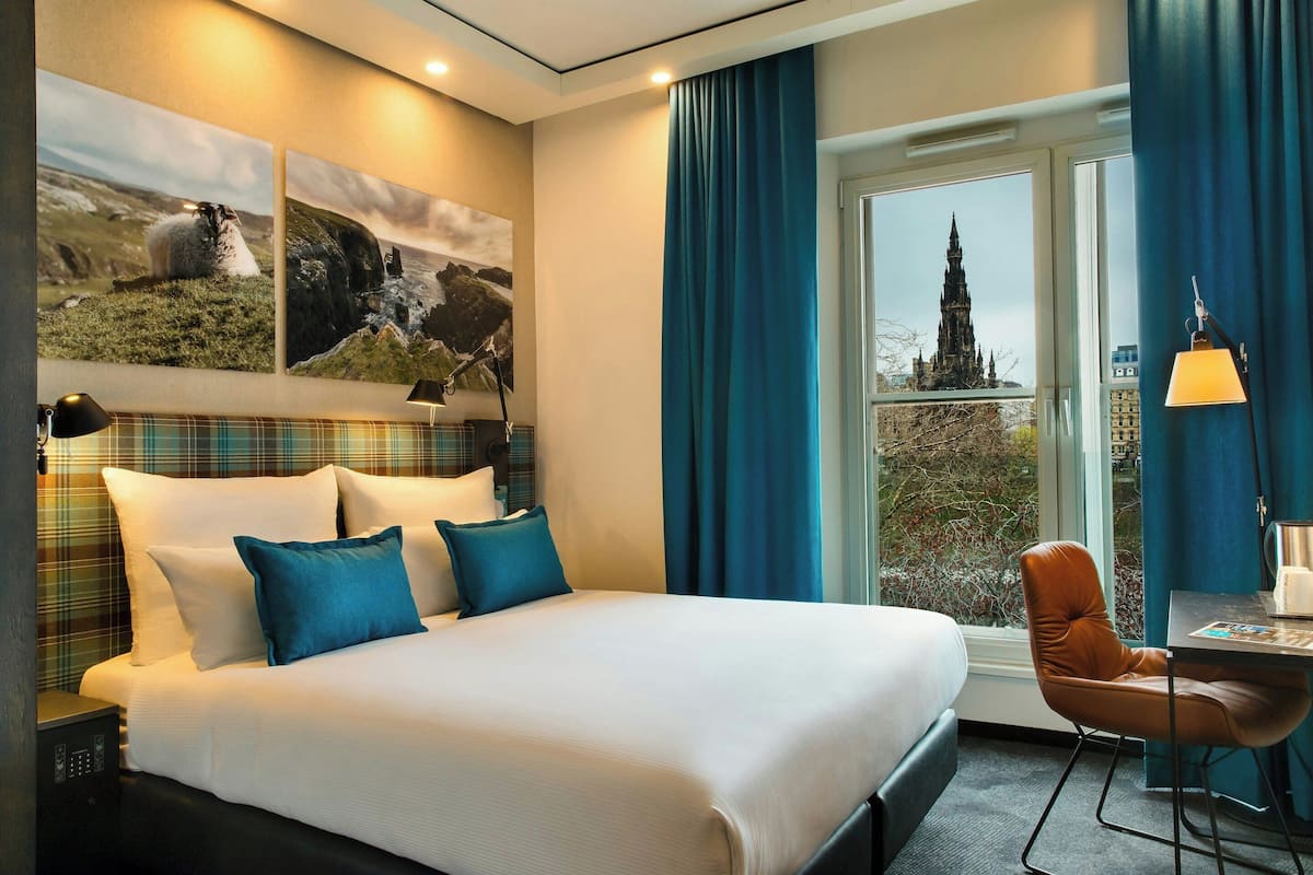 A hotel room with a large bed, a desk with a chair, and a window with a view of a building.