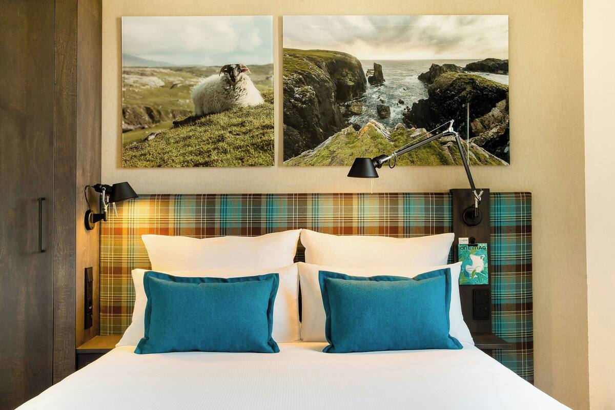 Une chambre d’hôtel avec un lit, deux oreillers, une table de chevet, une lampe et deux tableaux encadrés au mur.