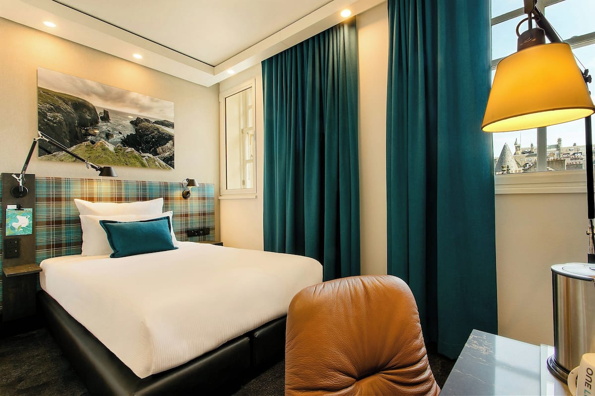 A hotel room with a bed, a desk, a chair, a lamp, and a window with curtains.