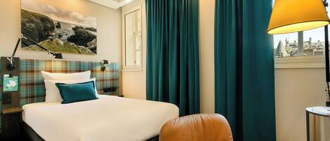 A hotel room with a bed, a desk, a chair, a lamp, and a window with curtains.