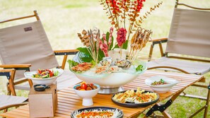 A wooden table with a variety of dishes, including a large bowl of seafood, salads, and other foods, set outdoors.