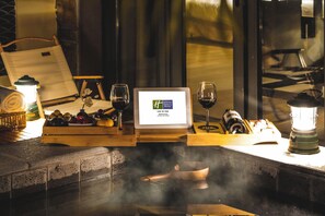 A laptop displaying a logo, a wooden table with a tray of fruits, two wine glasses, a bottle, and a lantern.