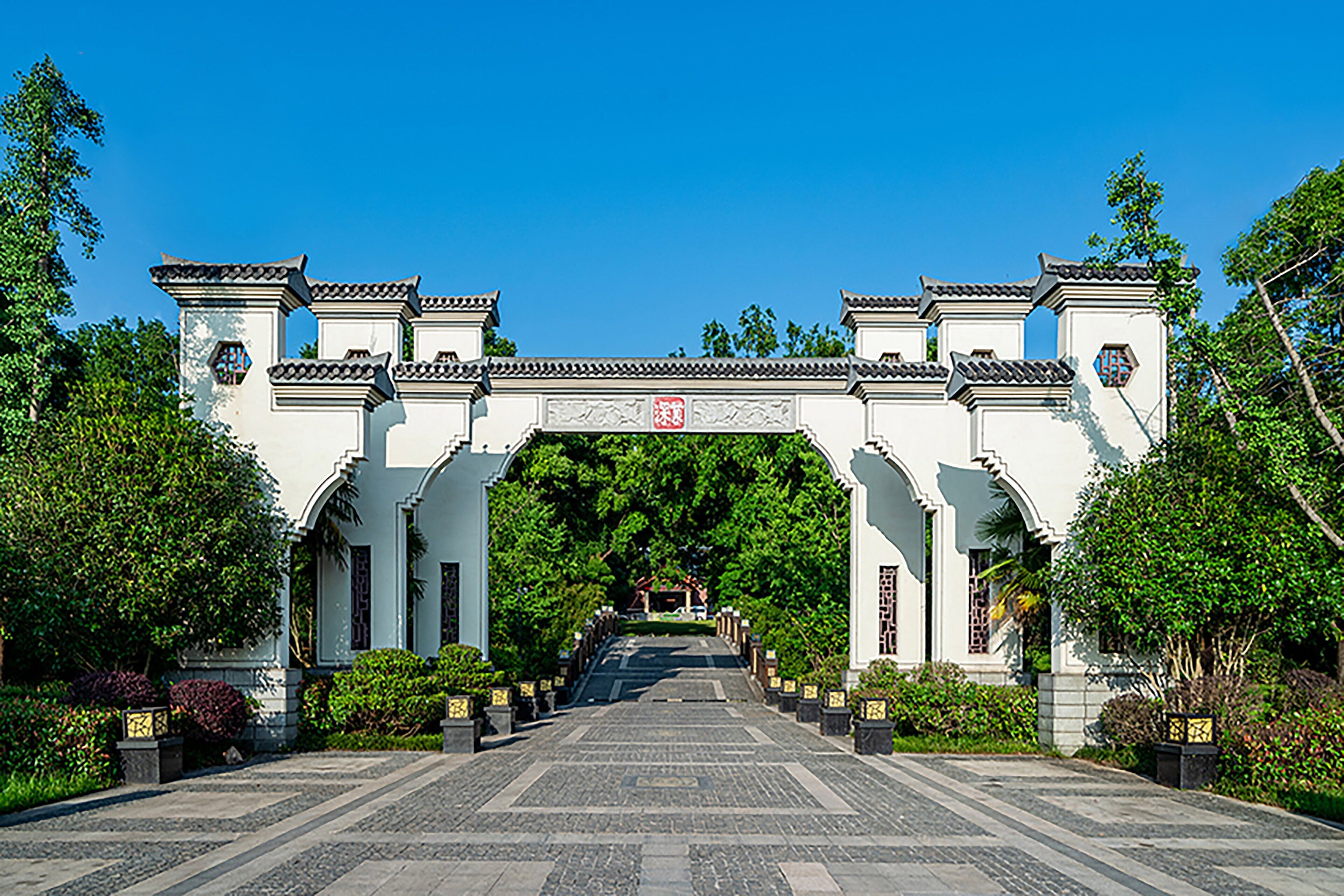A traditional gate with a red sign, flanked by white buildings and surrounded by greenery.