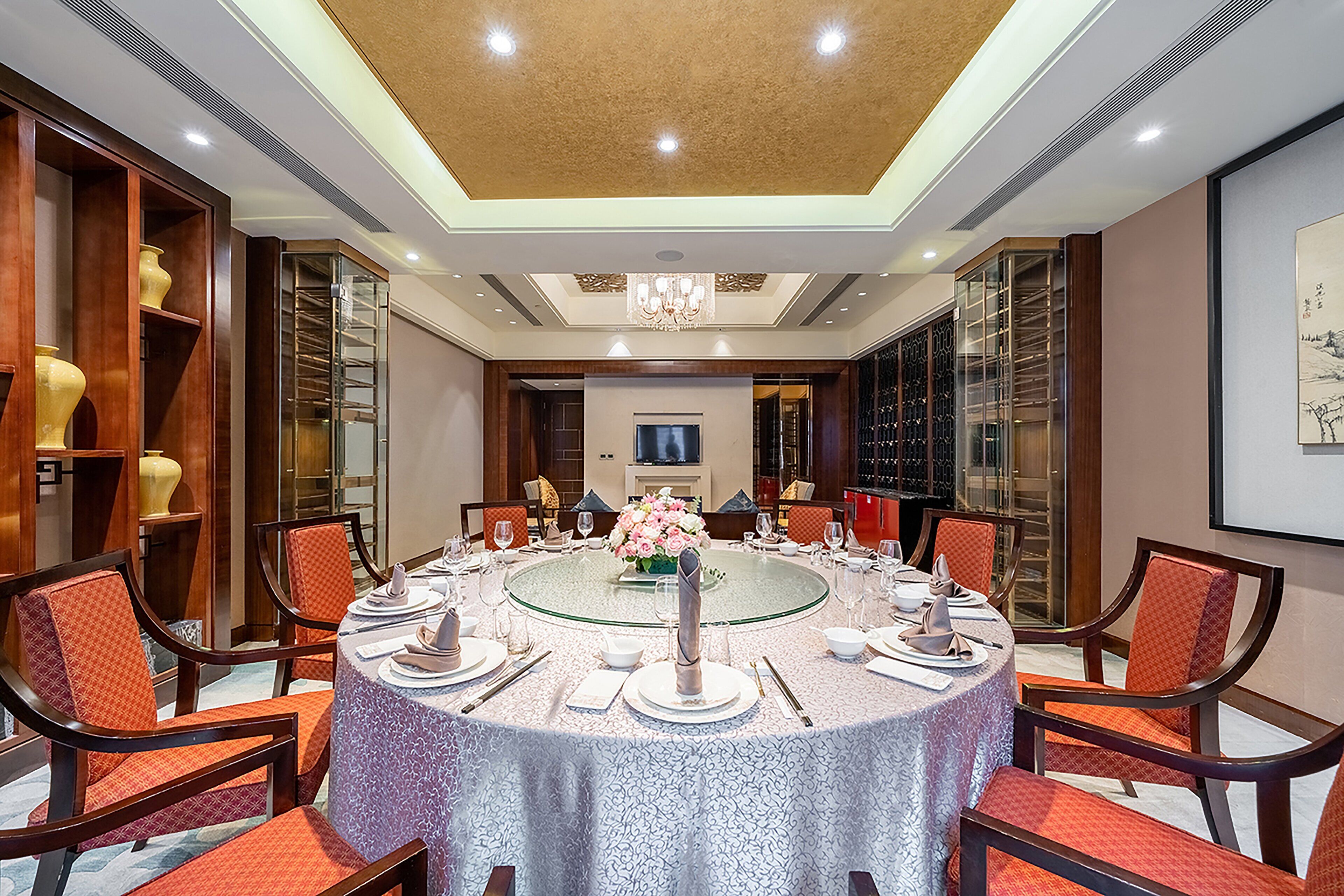 A conference room with a round table set for a meeting, surrounded by chairs, with a chandelier above and a large painting on the wall.