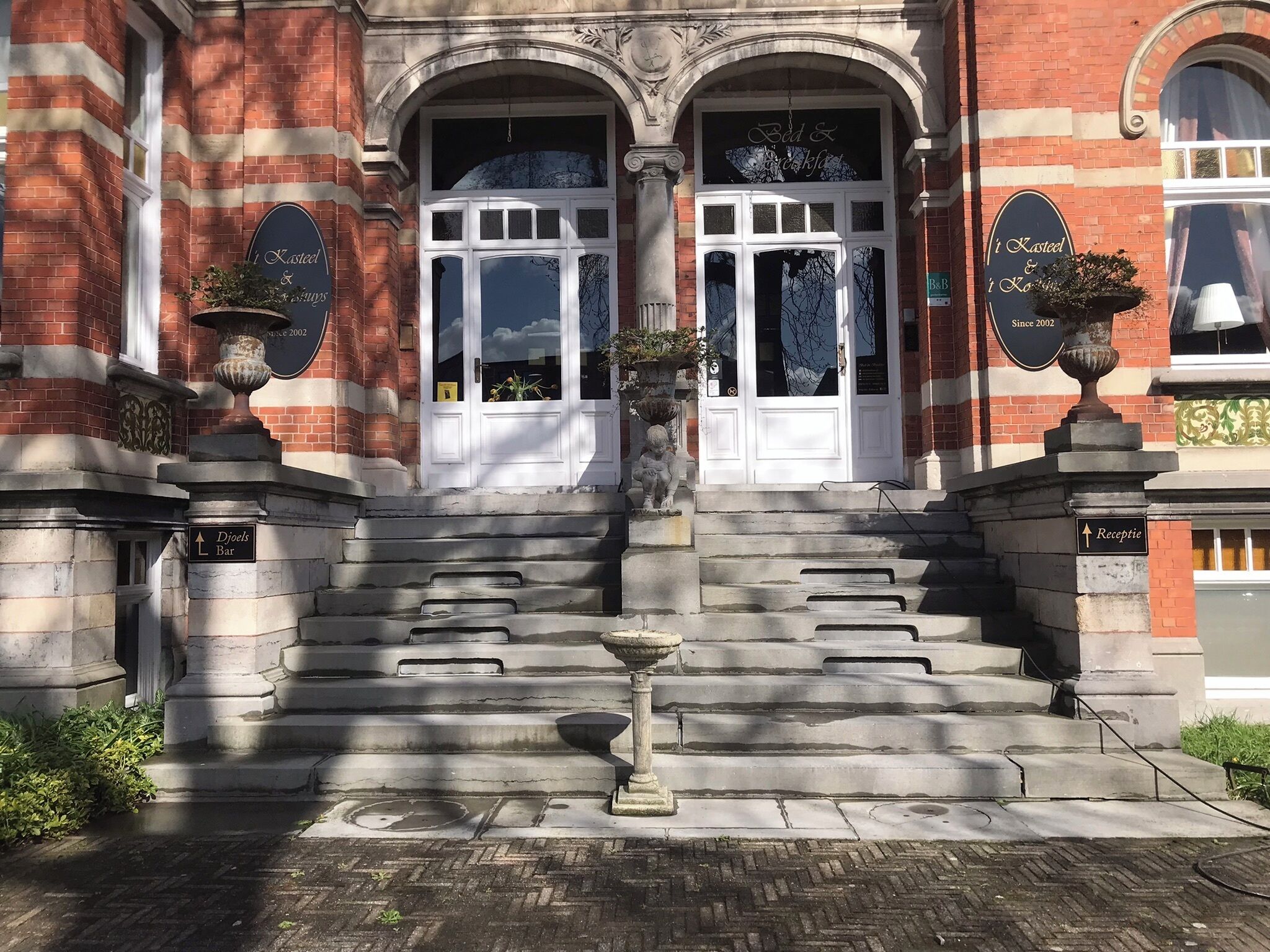 Een gebouw met een statig entree, stenen traptreden en aan weerszijden twee grote bloempotten.