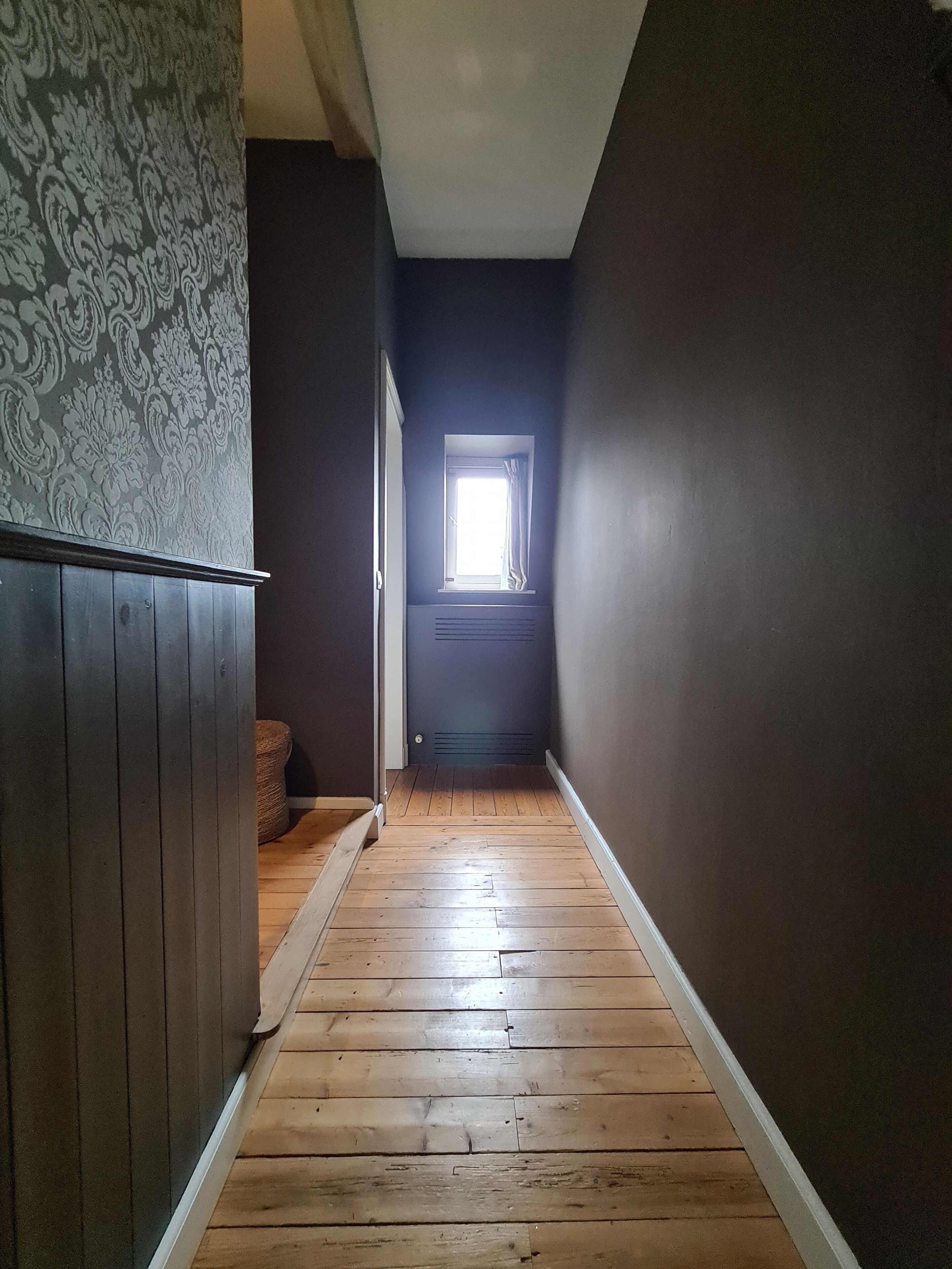 Un pasillo estrecho con suelo de madera, una ventana con cortinas y un papel tapiz estampado.