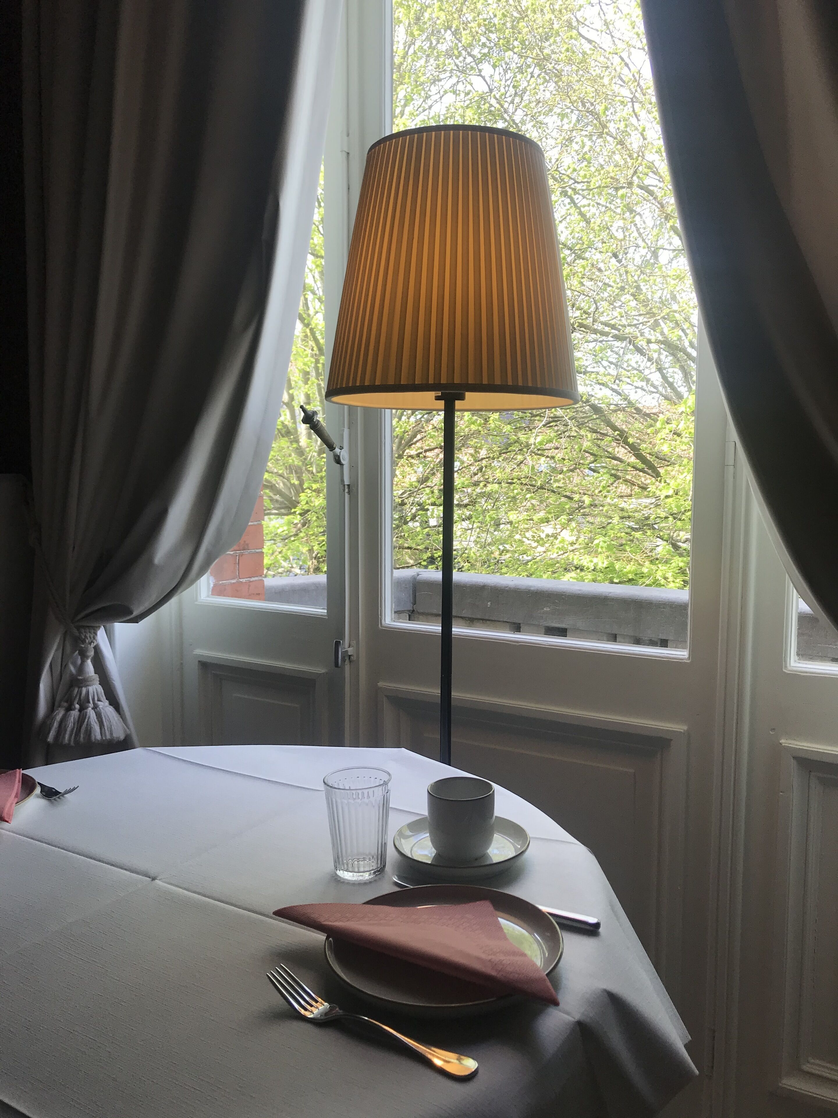 Een keurig gedekte eettafel met een lamp, een glas, een kopje, een bord, een servet en bestek.