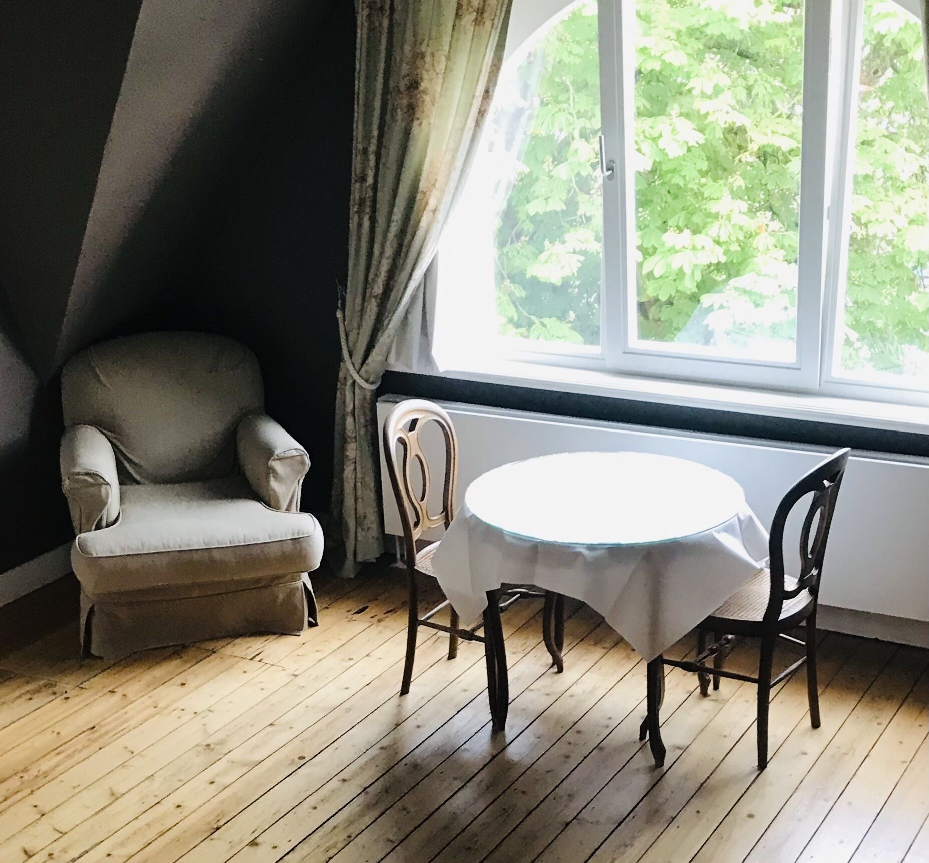 Une pièce dotée d’un plancher en bois, d’une table ronde recouverte d’une nappe blanche, de deux chaises, d’un fauteuil gris et d’une fenêtre donnant sur un feuillage verdoyant.