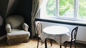 Une pièce dotée d’un plancher en bois, d’une table ronde recouverte d’une nappe blanche, de deux chaises, d’un fauteuil gris et d’une fenêtre donnant sur un feuillage verdoyant.