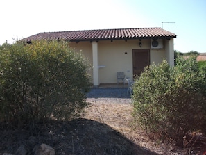 A small building with a tiled roof, a door, and a window. There is a chair outside and a gravel path leading to the entrance.