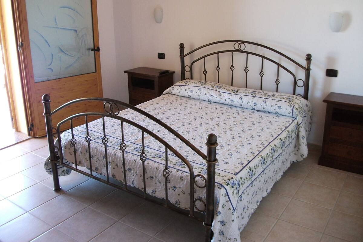 A single bed with a metal frame and floral bedding in a room with tiled flooring and a wooden door.