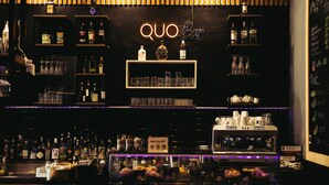A bar with shelves of liquor bottles, glasses, and a neon sign reading "QUO Bar". - QUO Milano - Hostel (Milan)