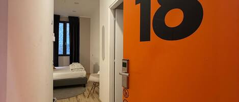 Un pasillo con piso de madera que conduce a una habitación con cama y ventana. Se ve una pared naranja con el número 18.