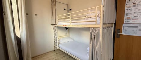 A bunk bed room with a wooden floor, a door with a notice board, and a window with curtains.