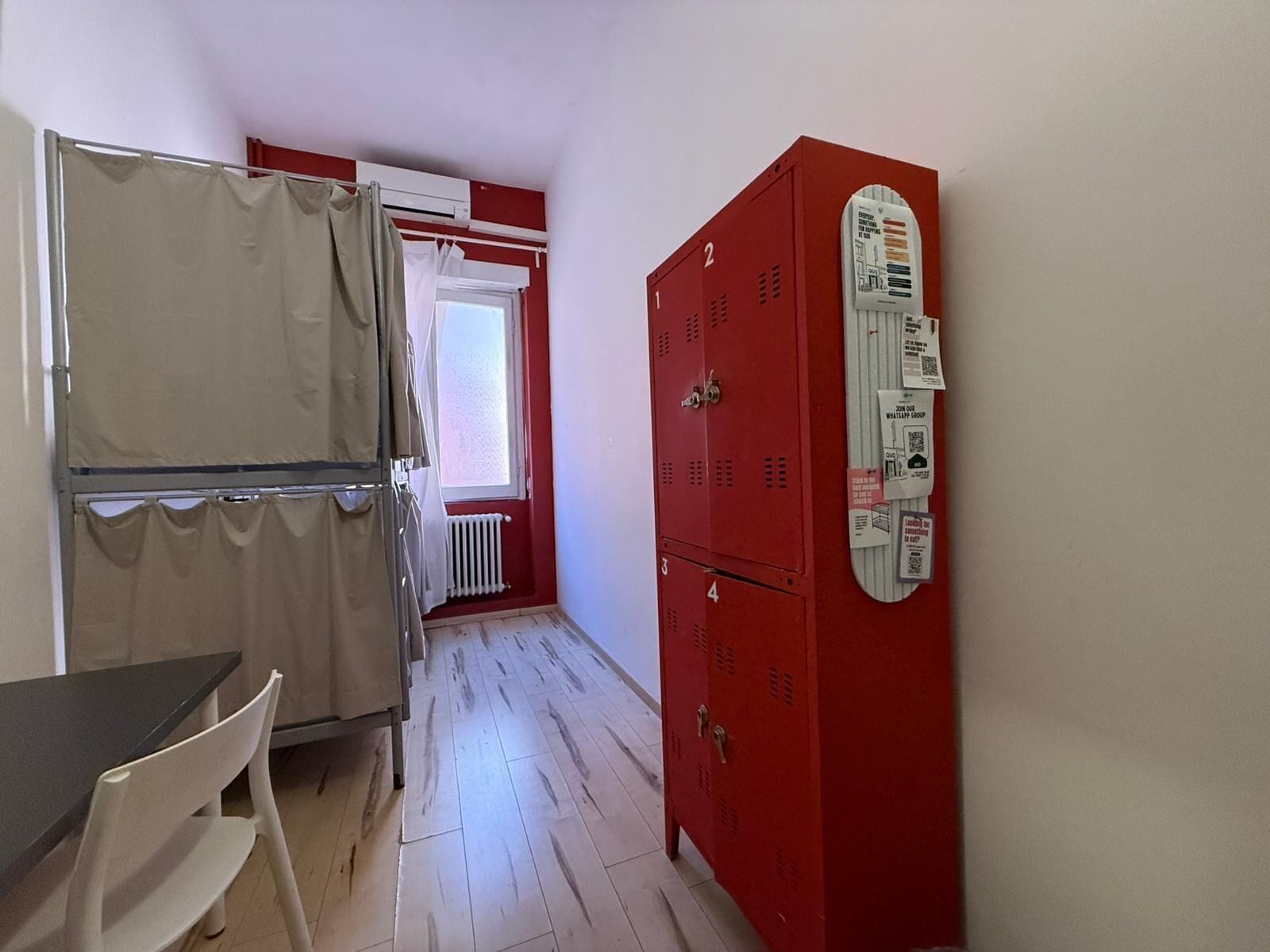 Ein Zimmer mit Tisch und Stühlen, einem Fenster mit Vorhängen und einem roten Funktionsschrank mit Bedienfeld.