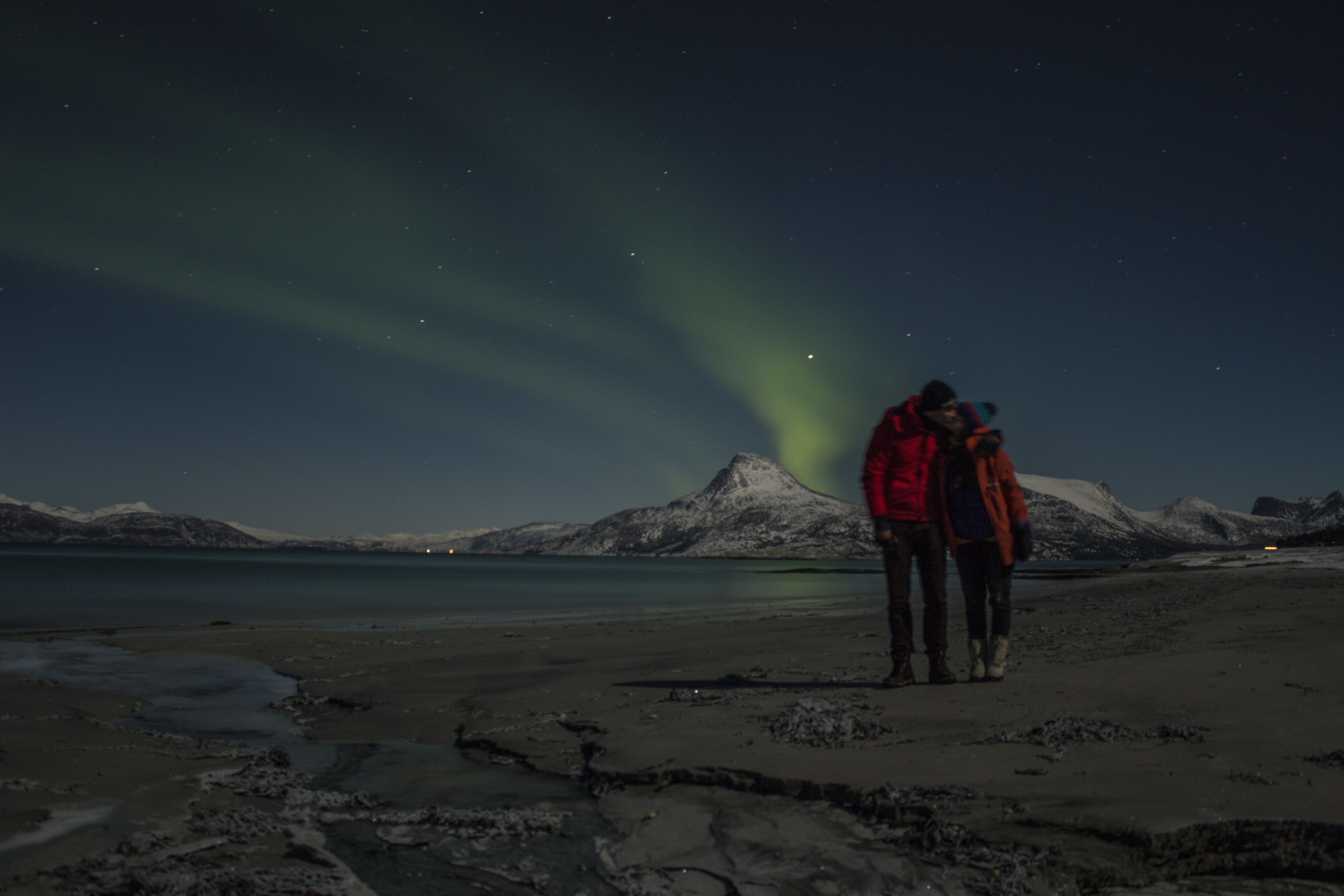 Un couple se tient sur une plage, avec une montagne en arrière-plan et des aurores boréales dans le ciel.
