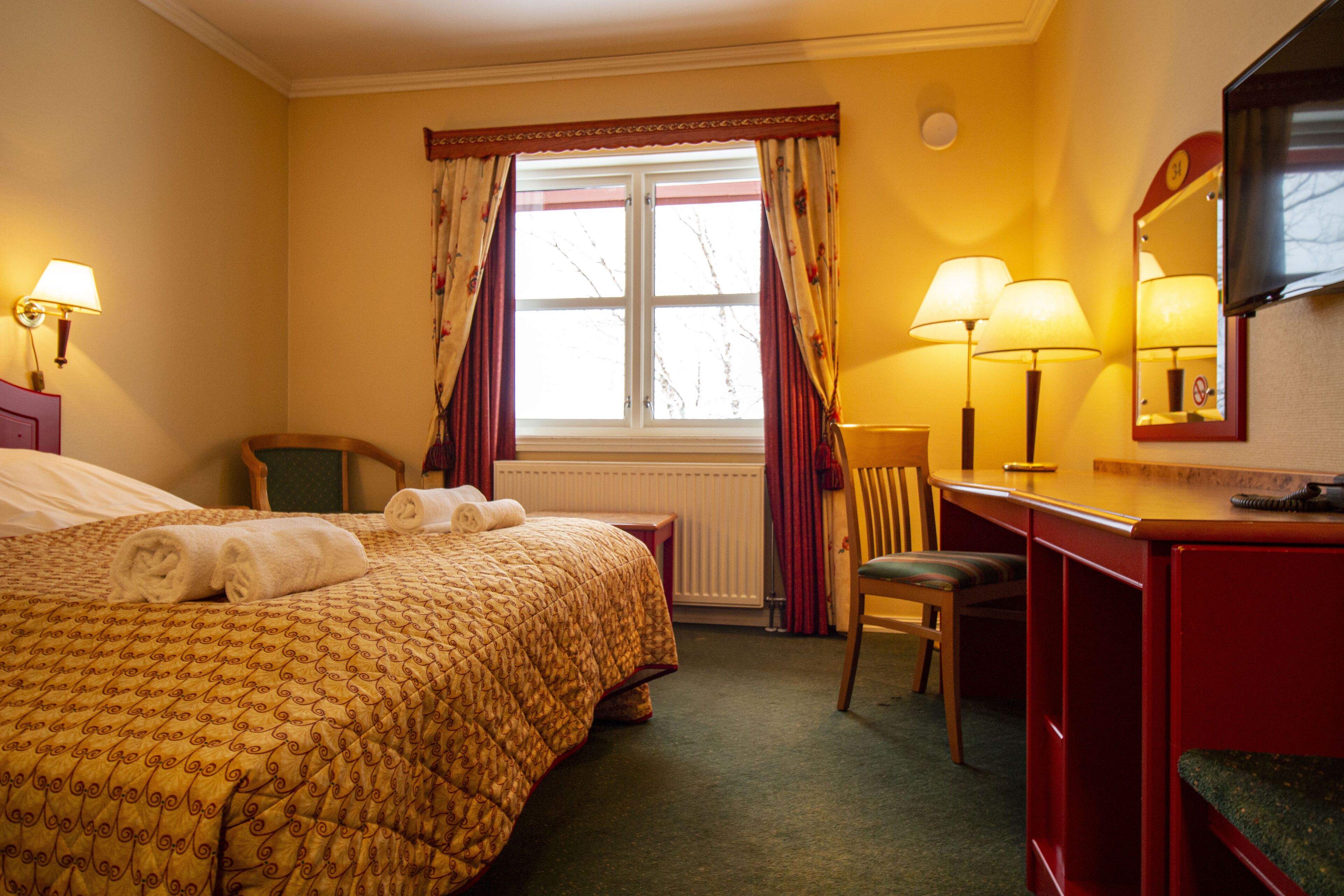 A hotel room with a bed, a desk, a chair, a window with curtains, and a mirror.