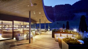 A modern outdoor lounge area with wooden flooring, seating arrangements, and a view of a mountainous landscape.