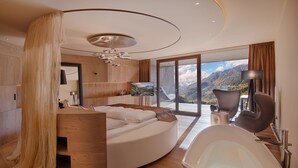 A modern bedroom with a curved bed, a large window with a mountain view, a bathtub, and a wooden floor.