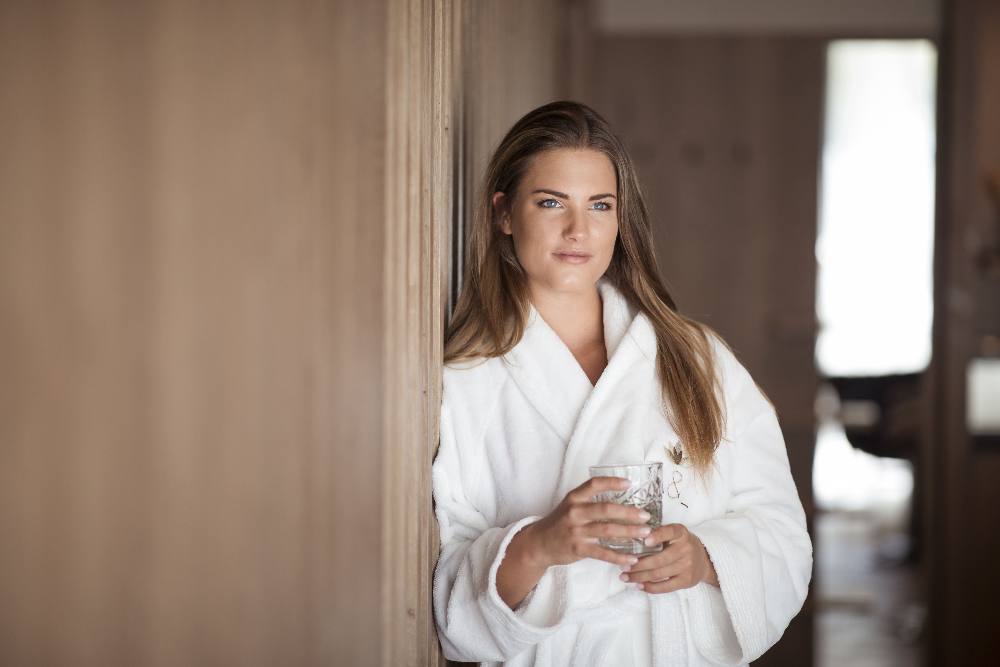 A person in a white robe holding a glass, standing in a hallway with wooden walls and a window in the background.