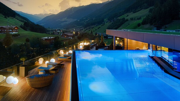 A modern hotel with a rooftop infinity pool, wooden deck seating, and a scenic mountain view.
