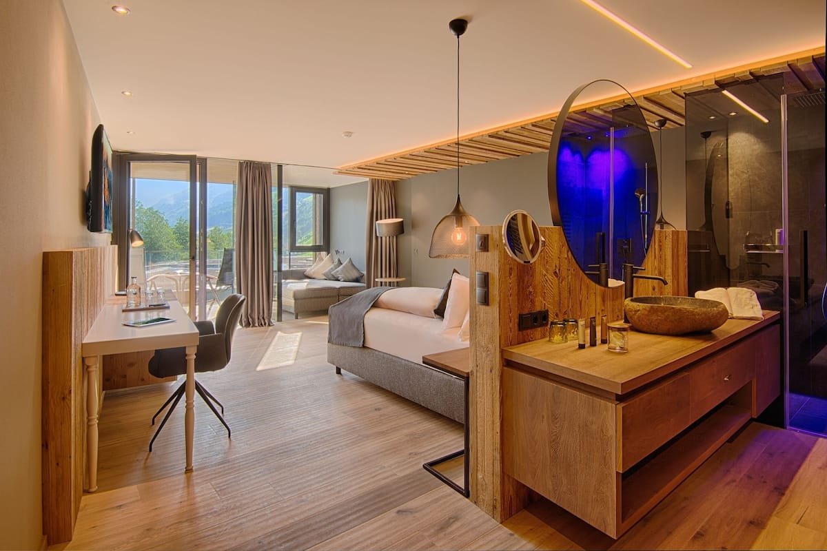 A modern hotel room with a wooden floor, a bed, a desk with a chair, a large mirror, and a bathroom with a glass shower.
