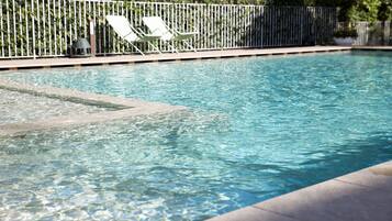 Una piscina con agua cristalina, rodeada por una valla blanca y dos sillas de descanso blancas.