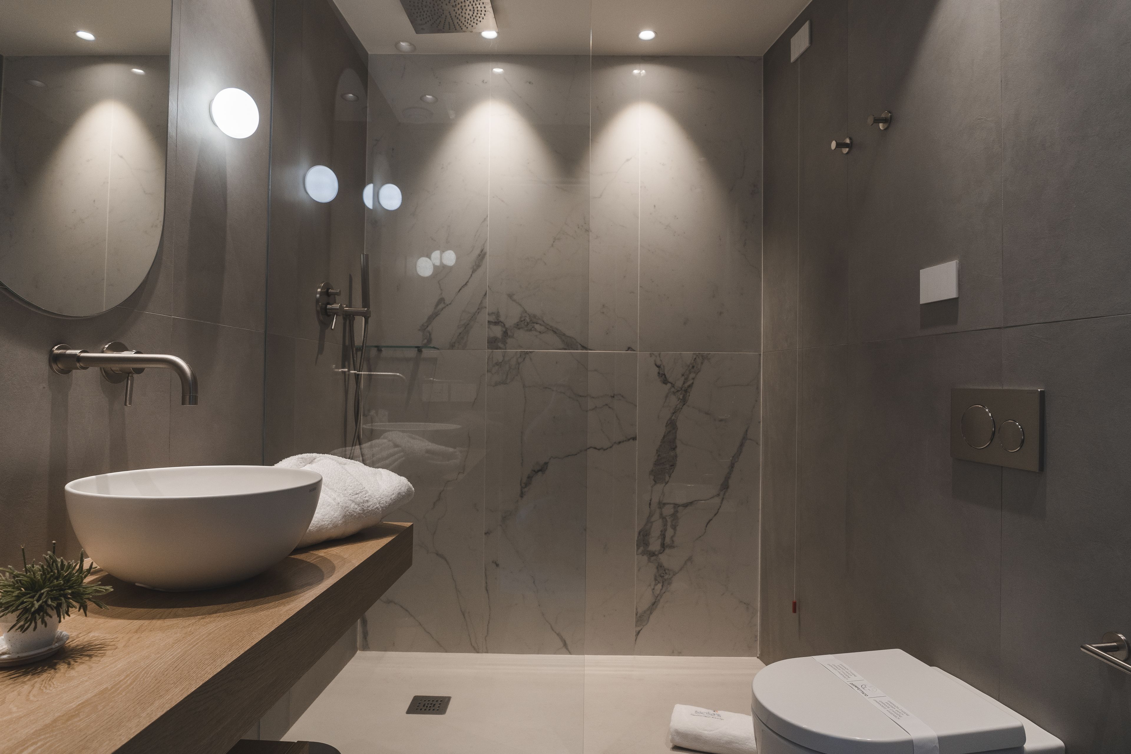 Un baño moderno con un lavamanos blanco de diseño, una encimera de madera, un espejo redondo y una zona de ducha con pared de mármol.