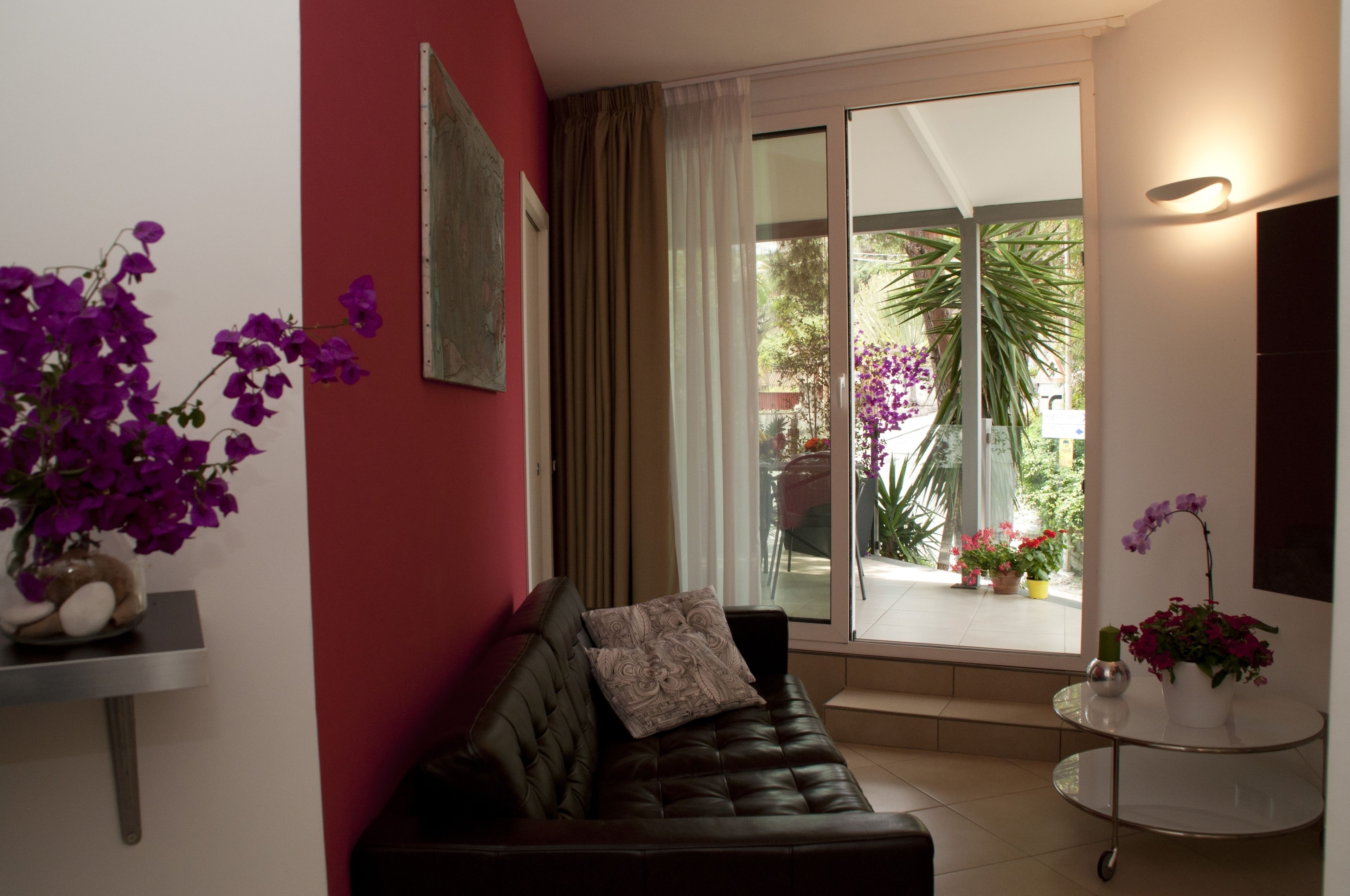 A living room with a dark brown sofa, a small table with a vase of purple flowers, and a large sliding glass door leading to a balcony with potted plants.