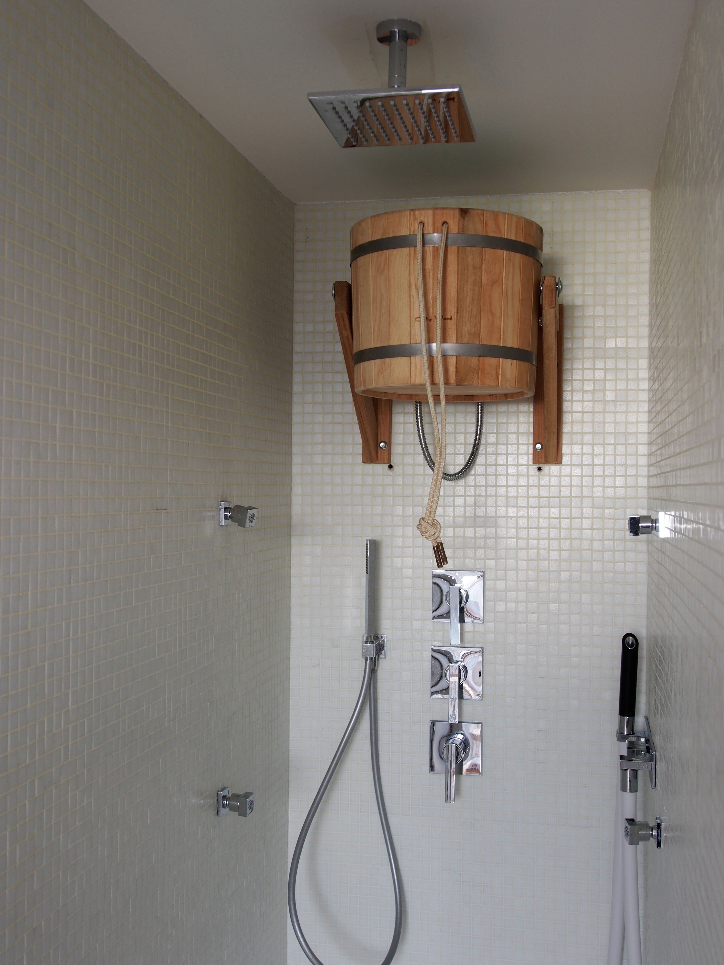 A shower area with a wooden bucket showerhead, a handheld showerhead, and a wall-mounted faucet.