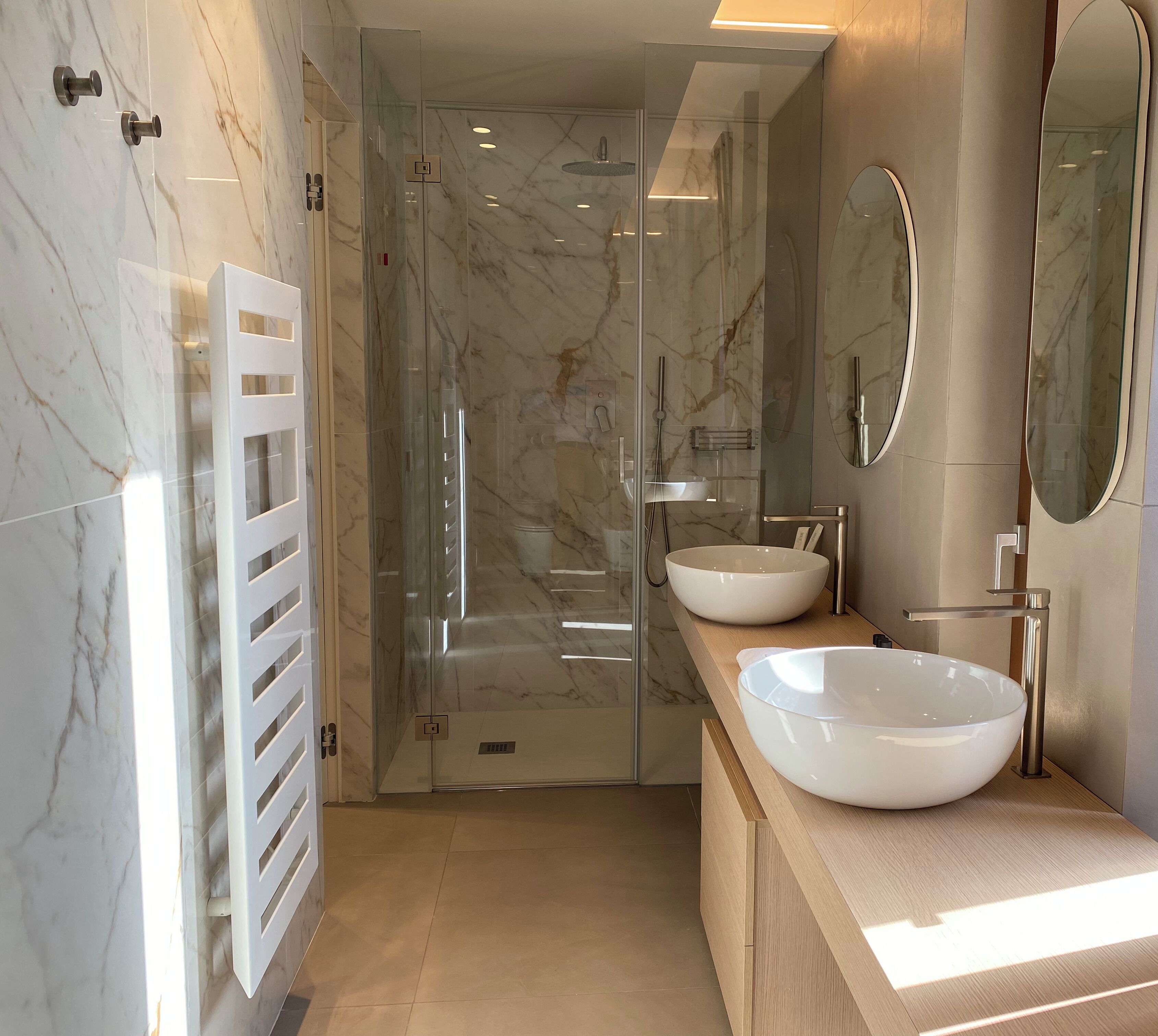 Une salle de bain moderne avec deux grands lavabos blancs, une douche à parois de verre et un miroir doté d’un cadre rond.