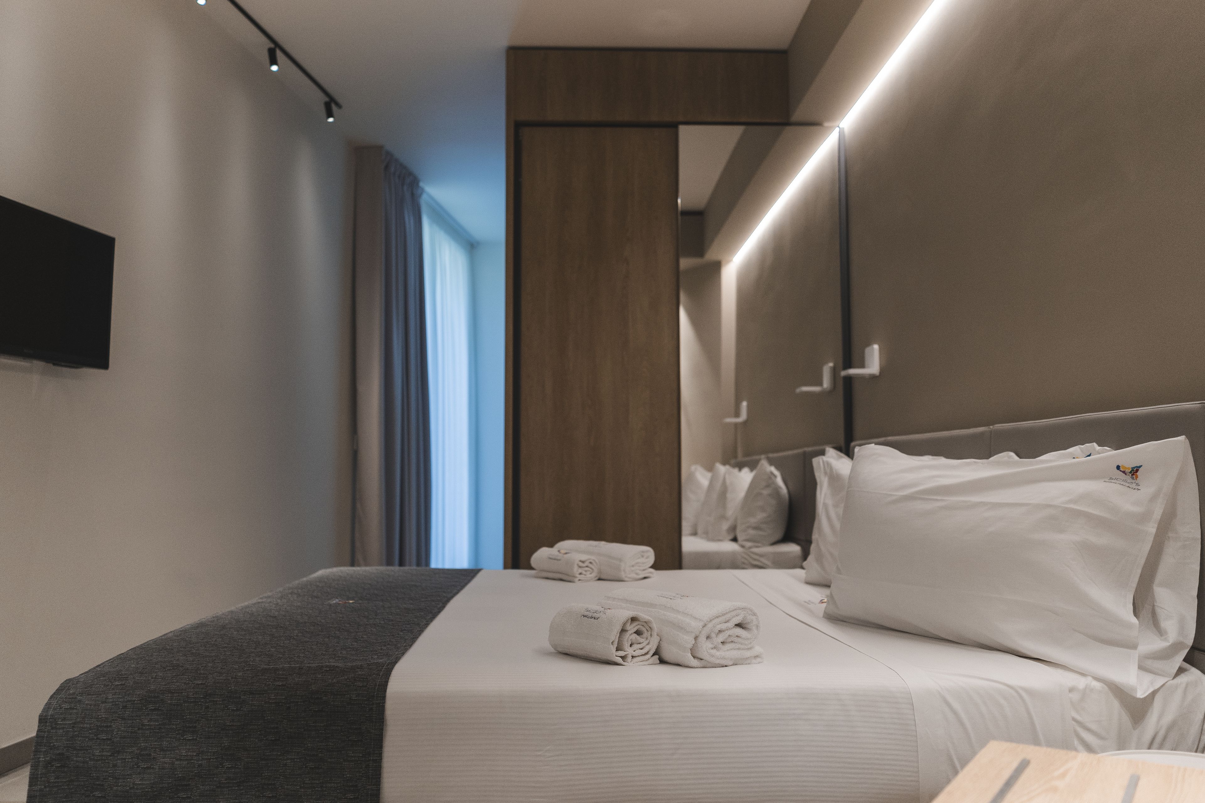 A modern hotel room with a neatly made bed, a flat-screen TV mounted on the wall, and a large mirror reflecting the room.